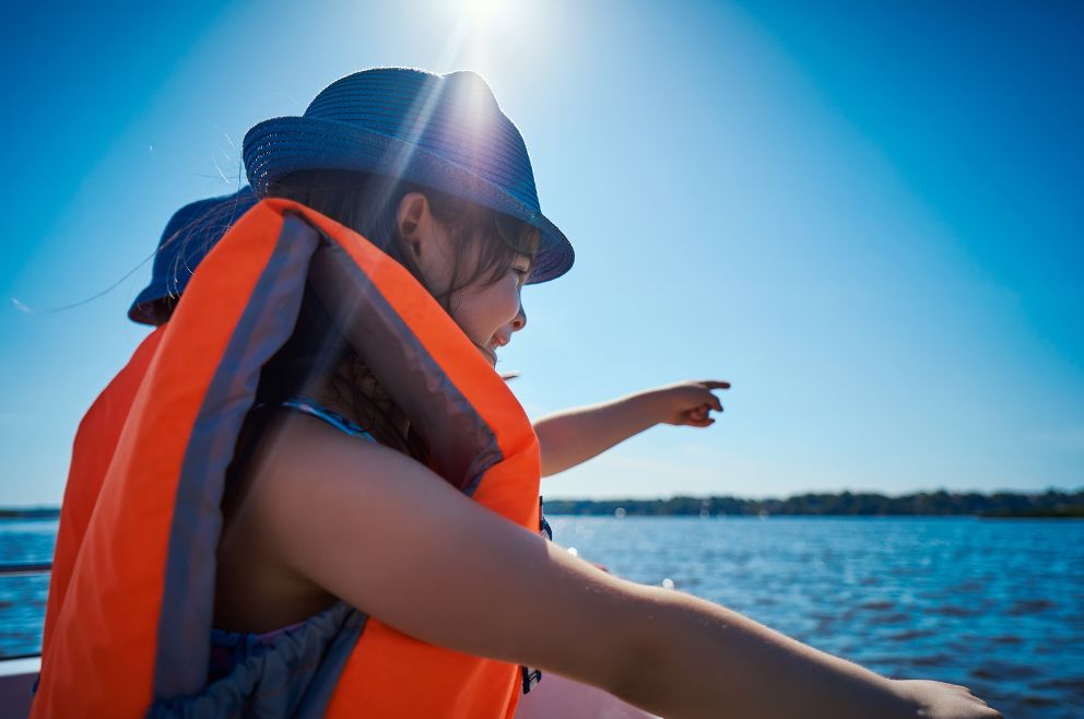 Image of a happy little girl looking and pointing at the water while wearing a hat and life jacket.
