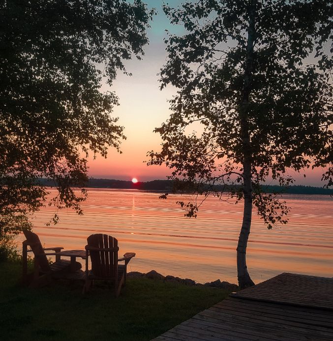 Landscape of sunset over the water two chairs and a dock silhouetted showing homes in Lac du Bonnet concept