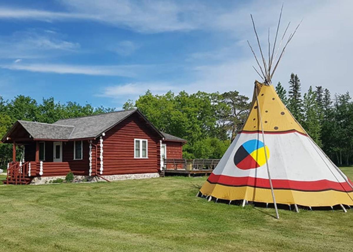 Photo of the Lac du Bonnet Museum beside a tipi. The Museum is a red log cabin.