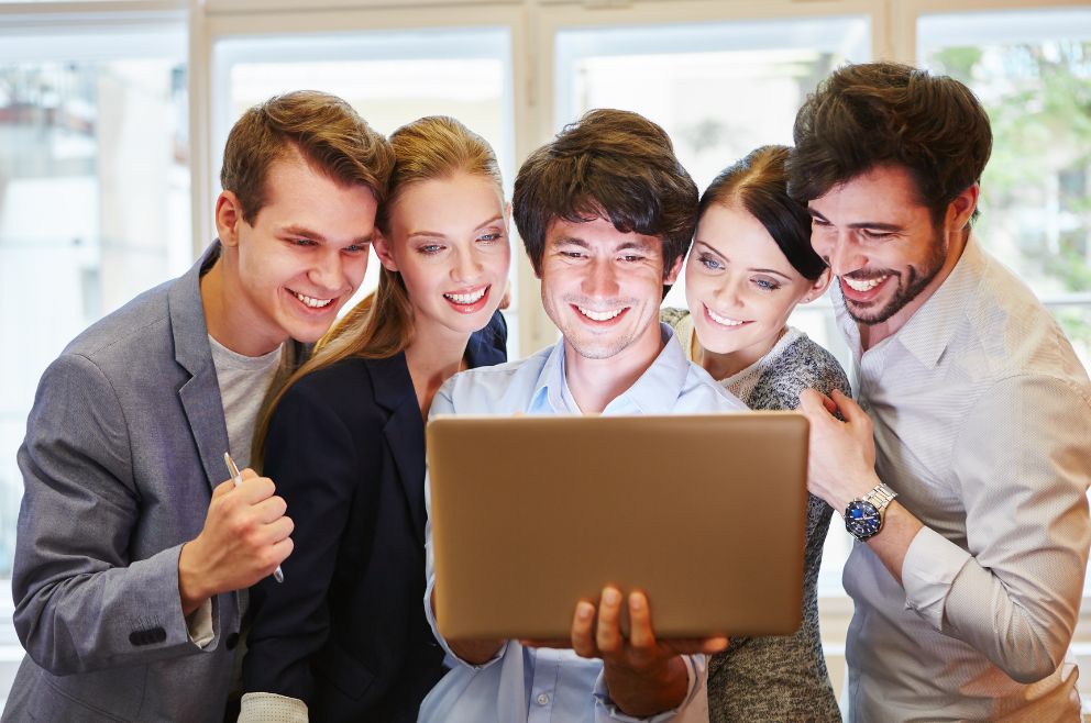 Photo of five happy people huddled together looking at a laptop.