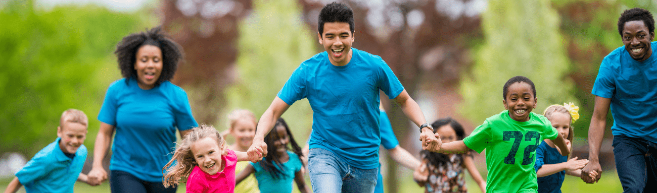 Photo is used to promote the Summer Camp Leader job showing a group of children and three camp leaders running while holding hands.
